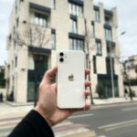 Close-up of a hand holding a smartphone on a street in Tirana, Albania, with modern buildings in the background.