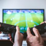 Close-up of hands holding gaming controllers in front of a TV. Engaged in a video gaming session.